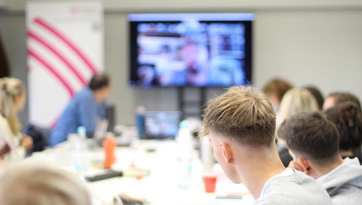 Eine Gruppe junger Menschen sitzt an einem langen Tisch, auf einem Bildschirm ist ein männlicher Referent per Zoom zugeschaltet