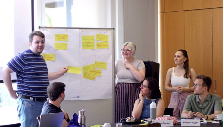 Ein junger Mann und zwei junge Frauen stehen vor einem Whiteboard und präsentieren ihre Ergebnisse auf Postits